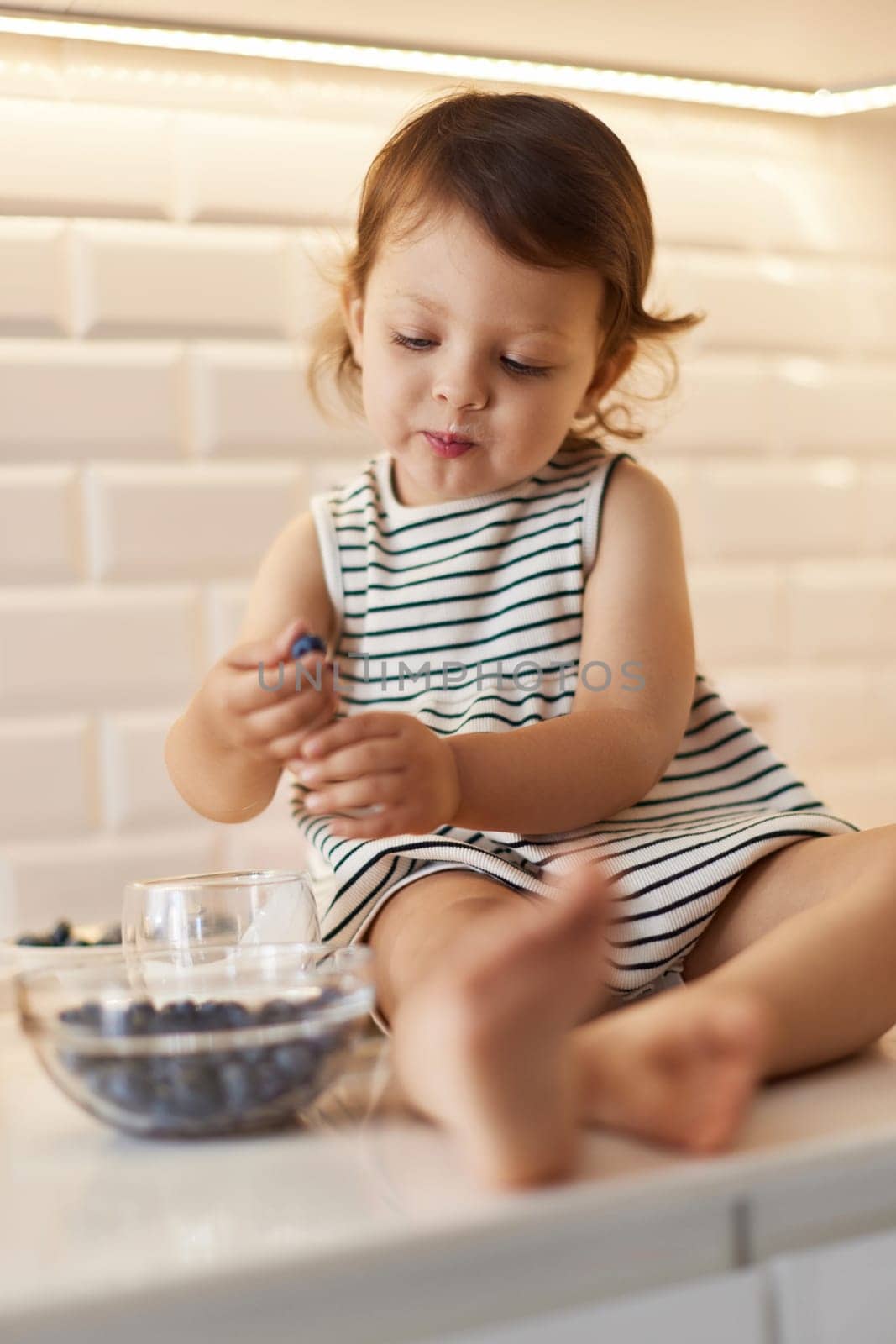 Little baby girl eating fresh blueberries from glass bowl in white kitchen