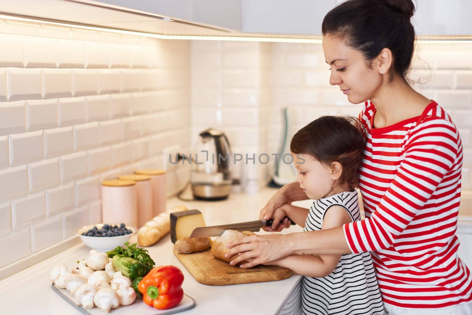 mother helps her cute little child daughter with knife for education in modern white kitchen