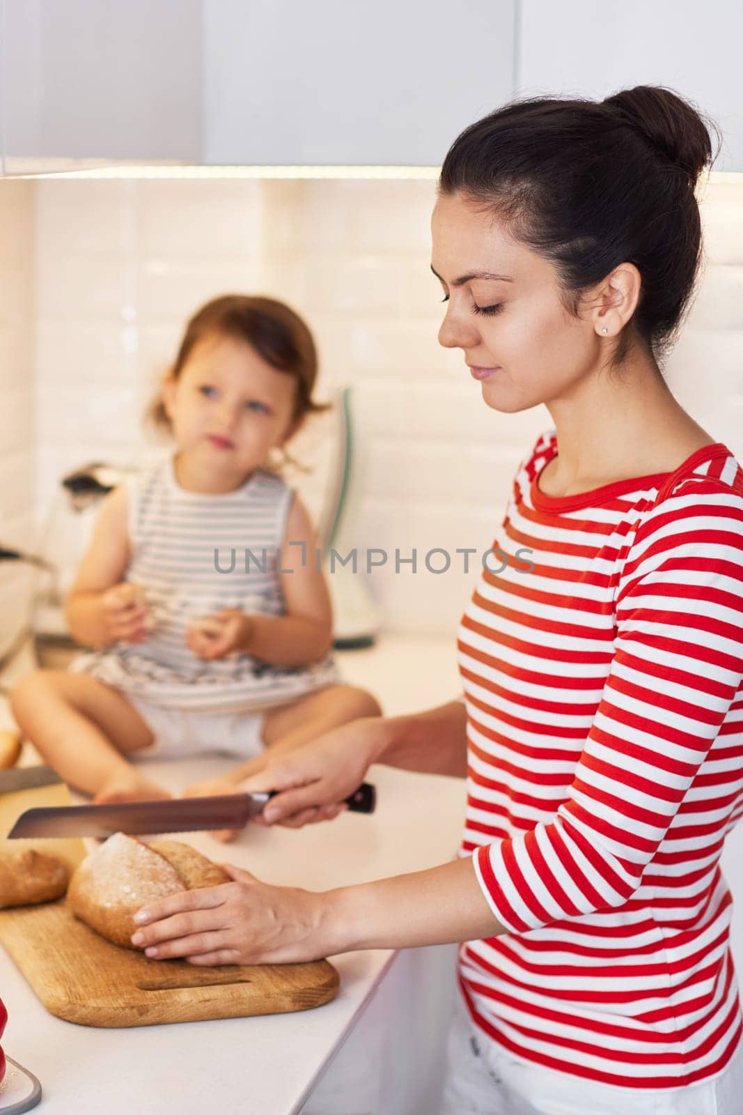 mother and cute little child daughter preparing breakfast in modern white kitchen. mom cutting bread on table.
