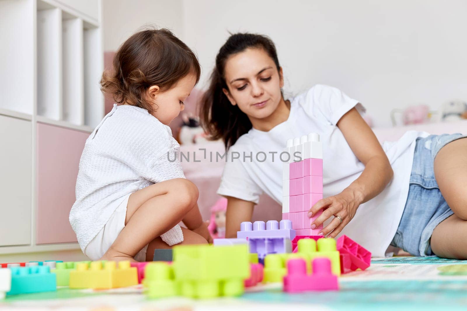 cute baby girl and her loving mother playing with colorful blocks in the children room, having fun together