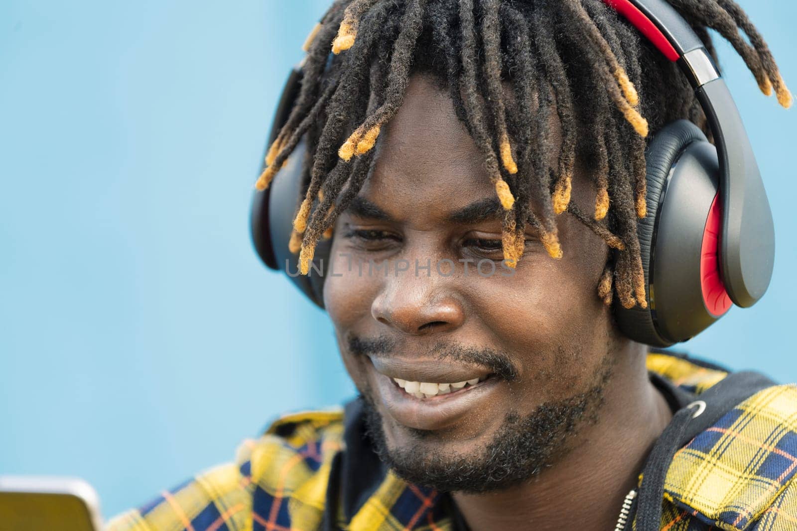 portrait of a young african man with headphones consulting his smart phone with a blue wall in the background, technology and lifestyle concept, copy space for text