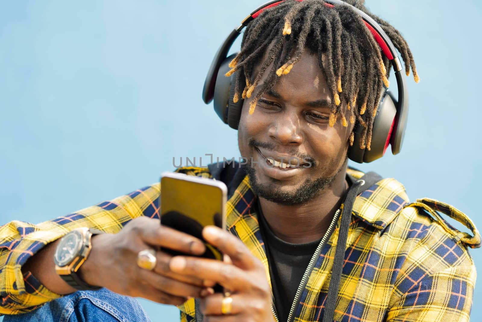 portrait of a young black man with a headphones writing on his smart phone with a blue wall in the background, technology and lifestyle concept, copy space for text