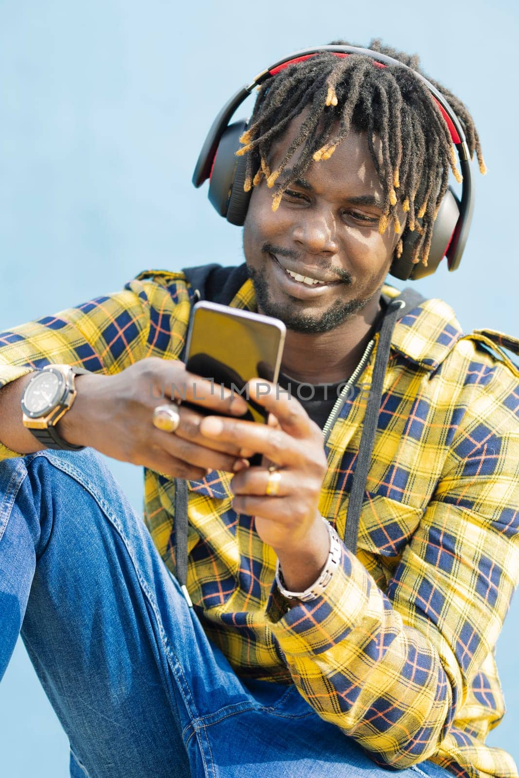 vertical portrait of a young black man with a headphones writing on his smart phone with a blue wall in the background, technology and lifestyle concept, copy space for text