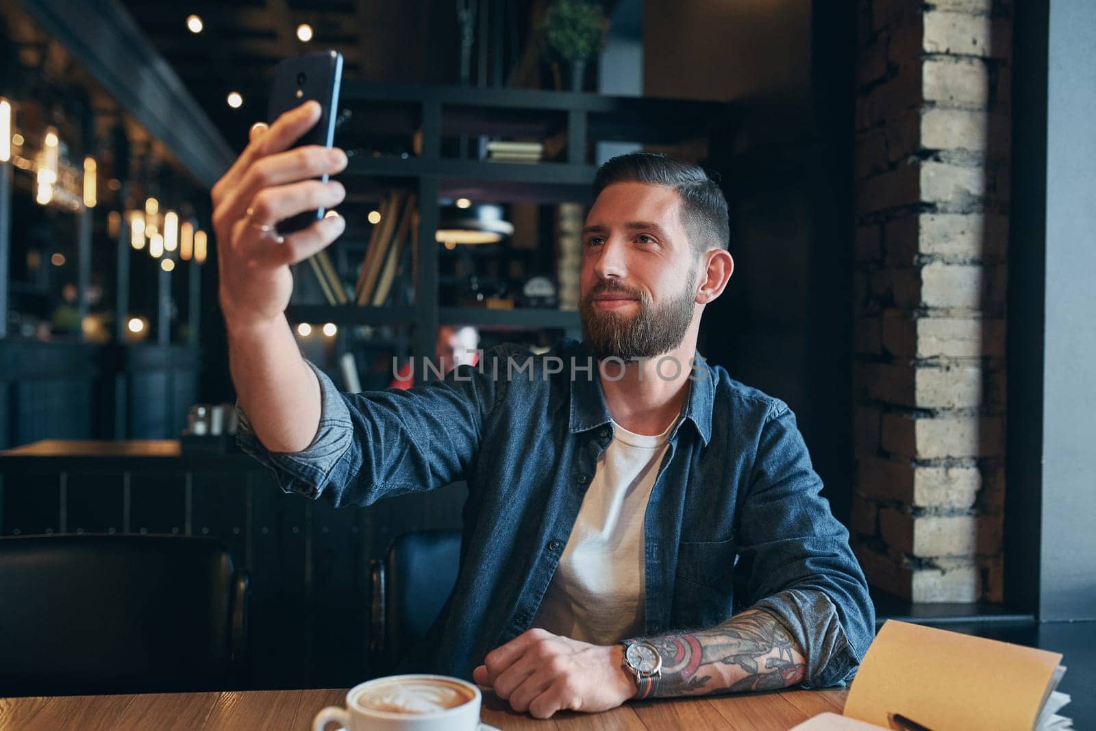 Man looking into the camera while making a selfie. Young bearded man, dressed in a denim shirt, sitting at table in cafe and use smartphone