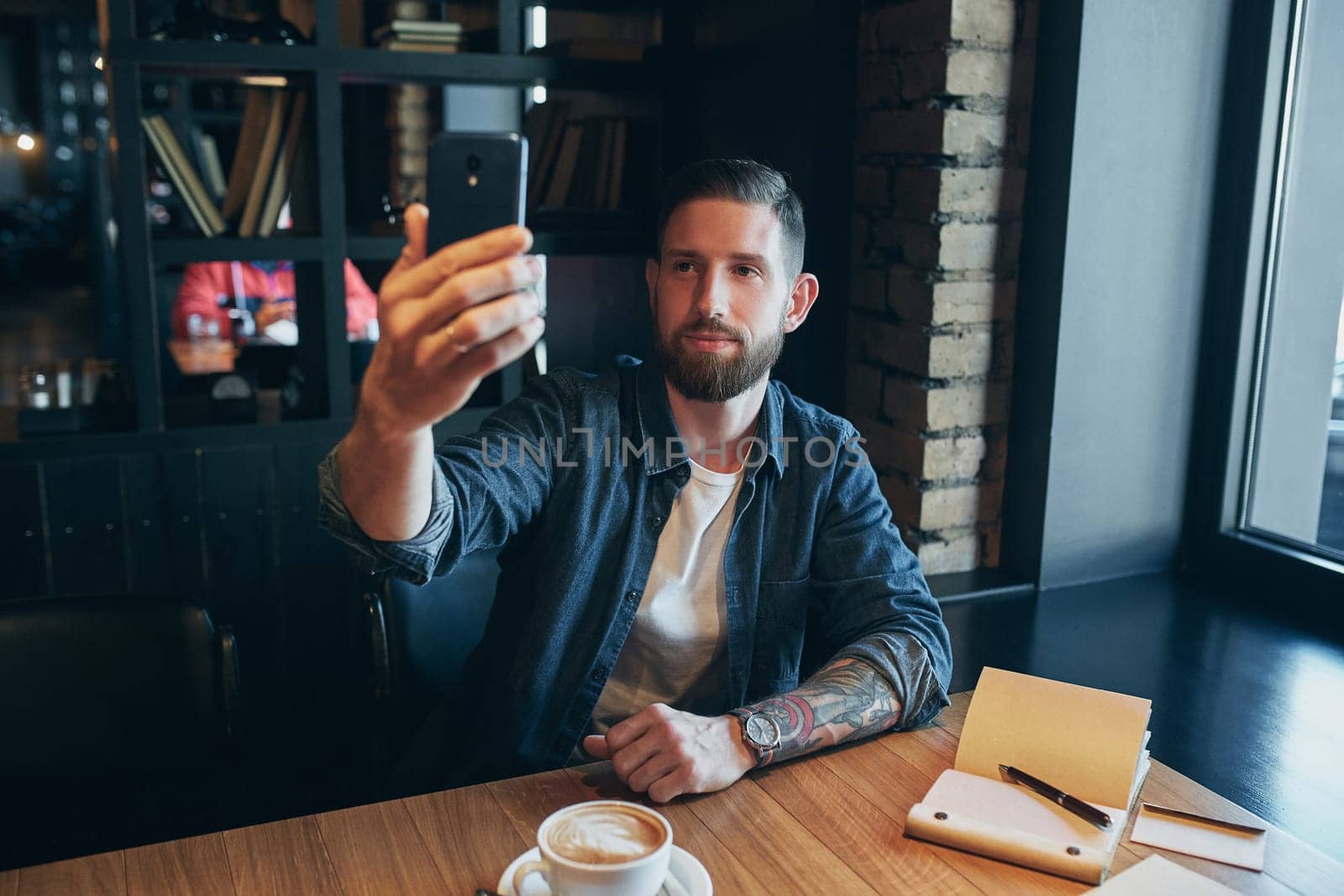 Man looking into the camera while making a selfie. Young bearded man, dressed in a denim shirt, sitting at table in cafe and use smartphone