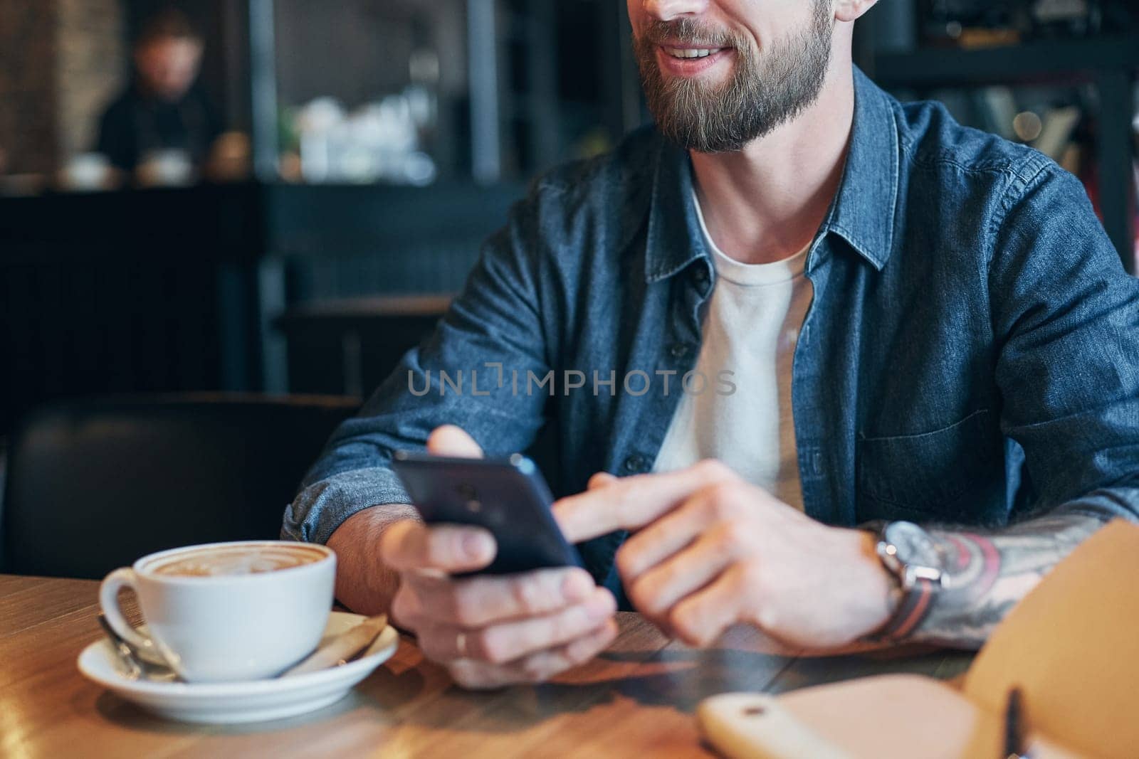 Man hands in denim shirt slide with finger on screen his smart phone, near cup with coffee on wooden table in cafe shop during his coffee break