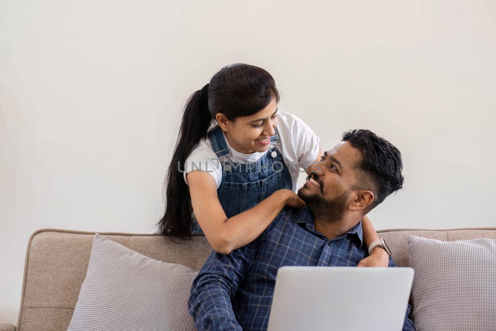 Cropped view of young woman hugging husband while using laptop on sofa in living room.