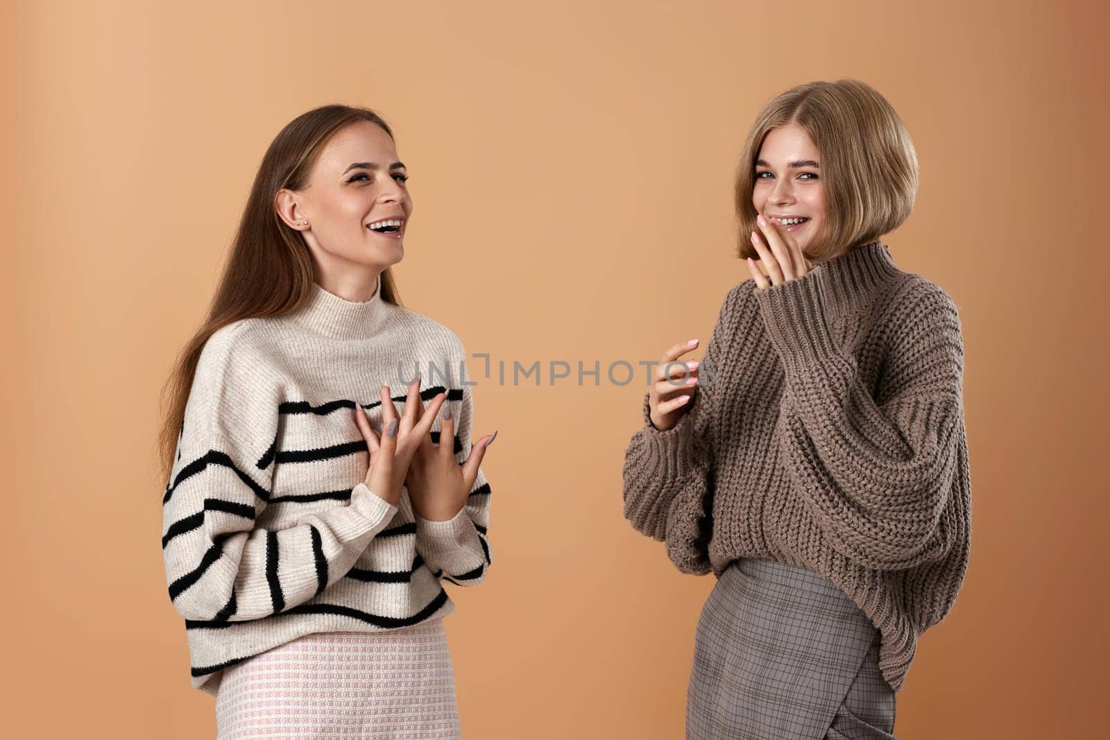 Two cheerful cheery women friends talking together while standing on beige background