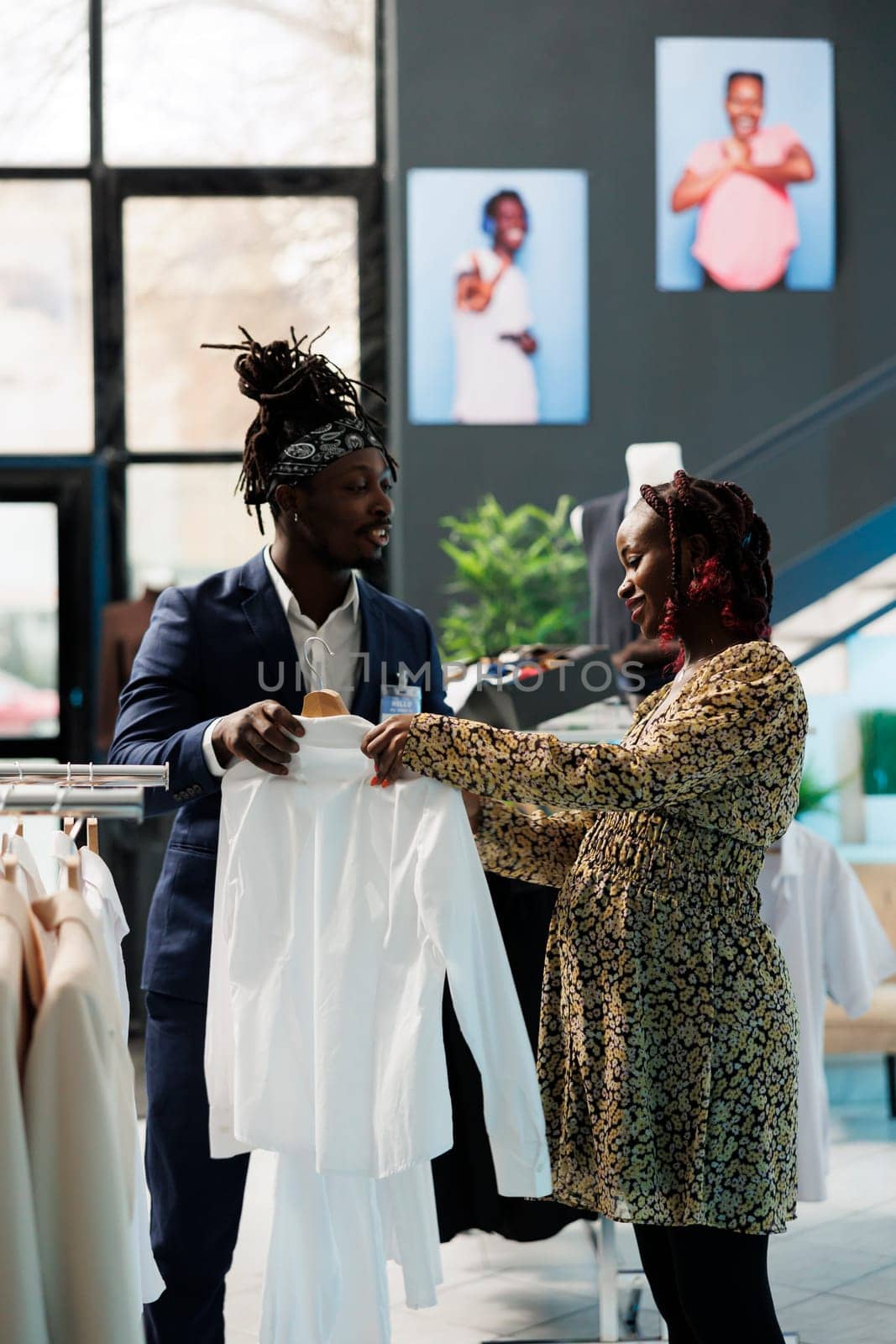 Showroom worker helping customer choosing white shirt, discussing clothes fabric in clothing store. African american pregnant woman buying fashionable maternity merchandise in modern boutique