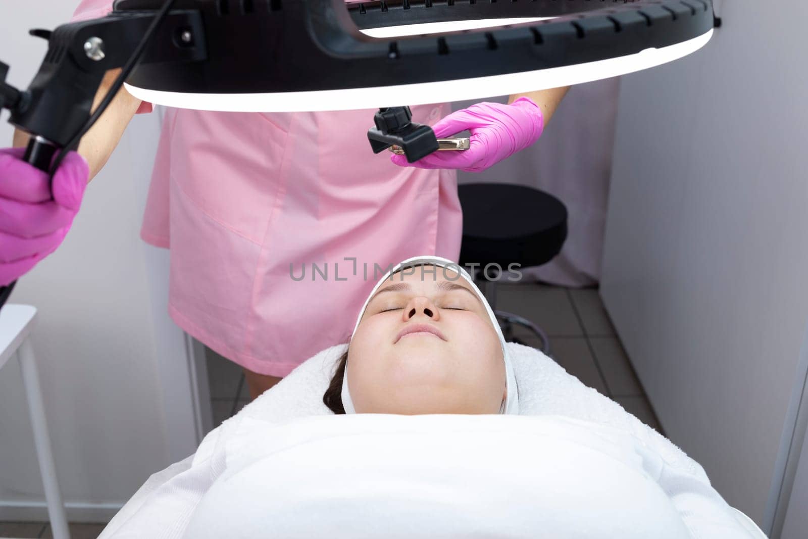 Cosmetologist Takes Picture of Female Patient On Phone. Young Pretty Woman Lying On Couch Below Led Lamp In Spa Salon. Beauty Procedure, Cosmetology, Wellness. Horizontal Plane. High quality photo