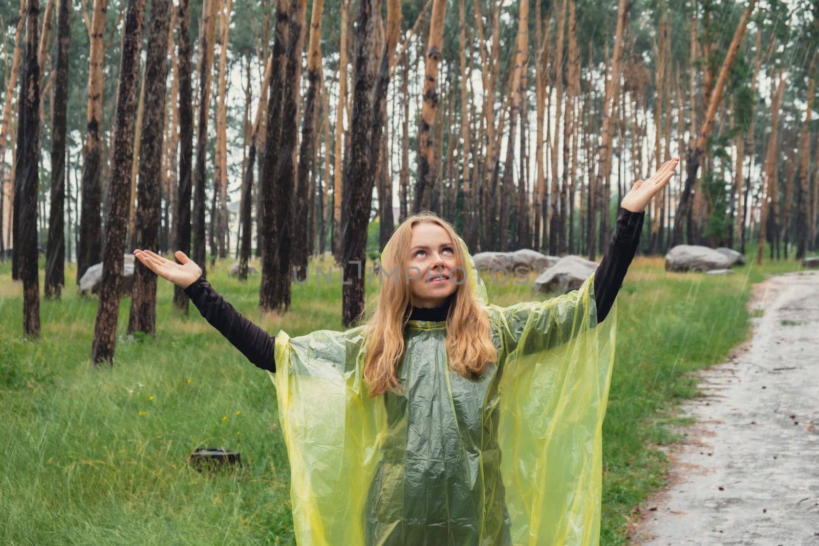 Happy blonde woman in yellow raincoat enjoying outdoors in forest. Rainy weather outside leisure activities in autumn time. Rainy season forecast