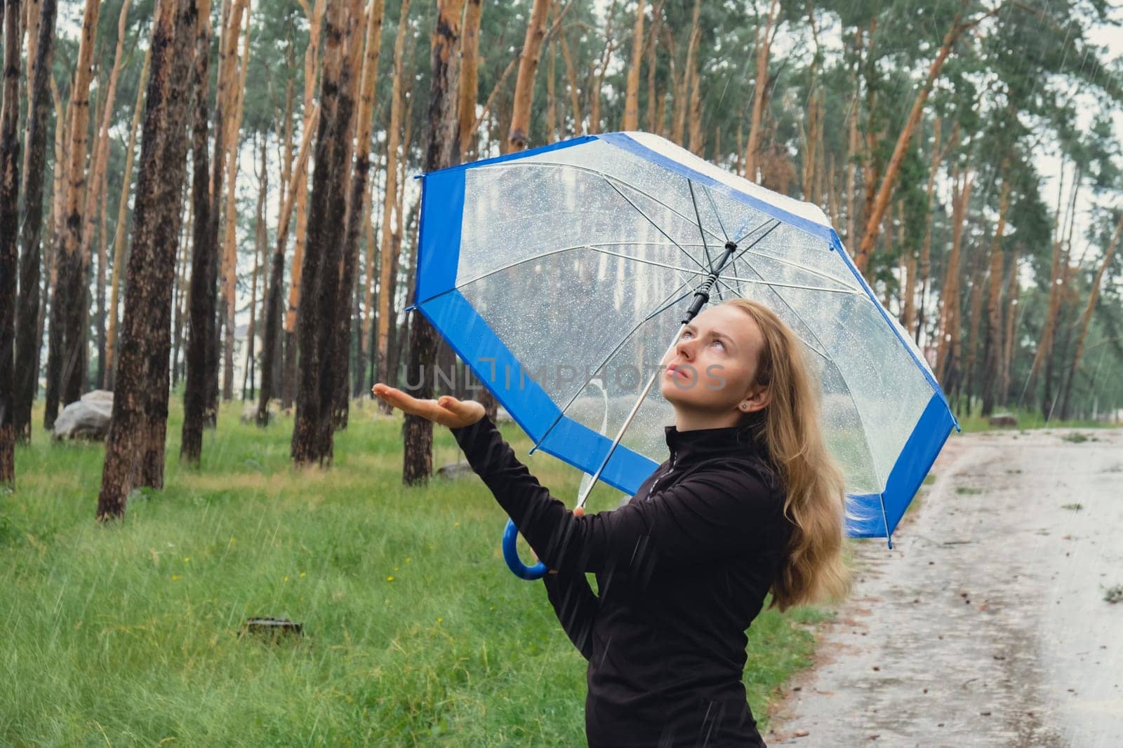 Young woman holding transparent blue umbrella outdoors in forest. Rainy weather day using umbrella. Woman with hand checking how long it will be raining by anna_stasiia