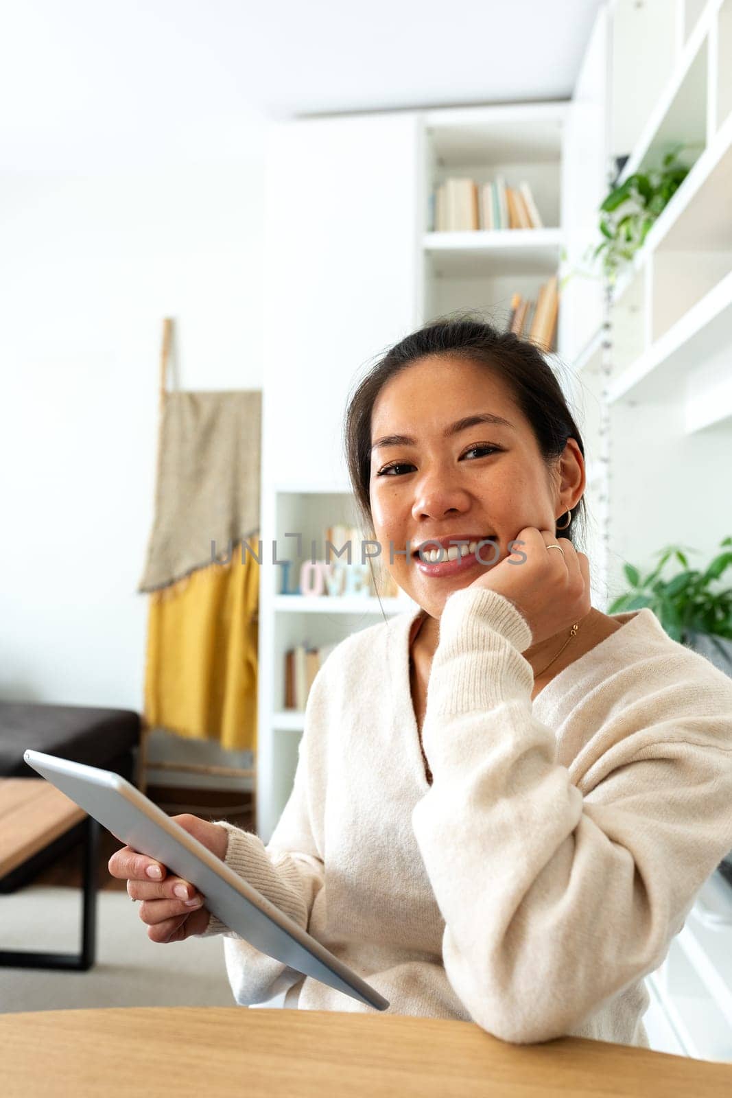 Happy young Asian woman working at home office holding tablet looking at camera. by Hoverstock