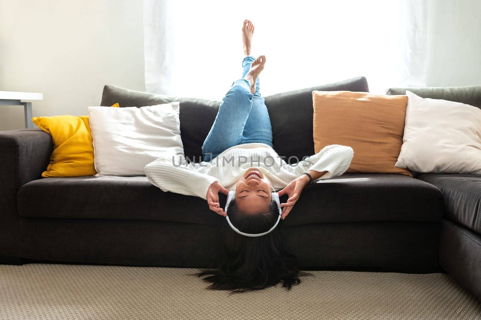 Happy young Asian woman relaxing at home, lying on the couch upside down, listening to music with headphones. Lifestyle concept.