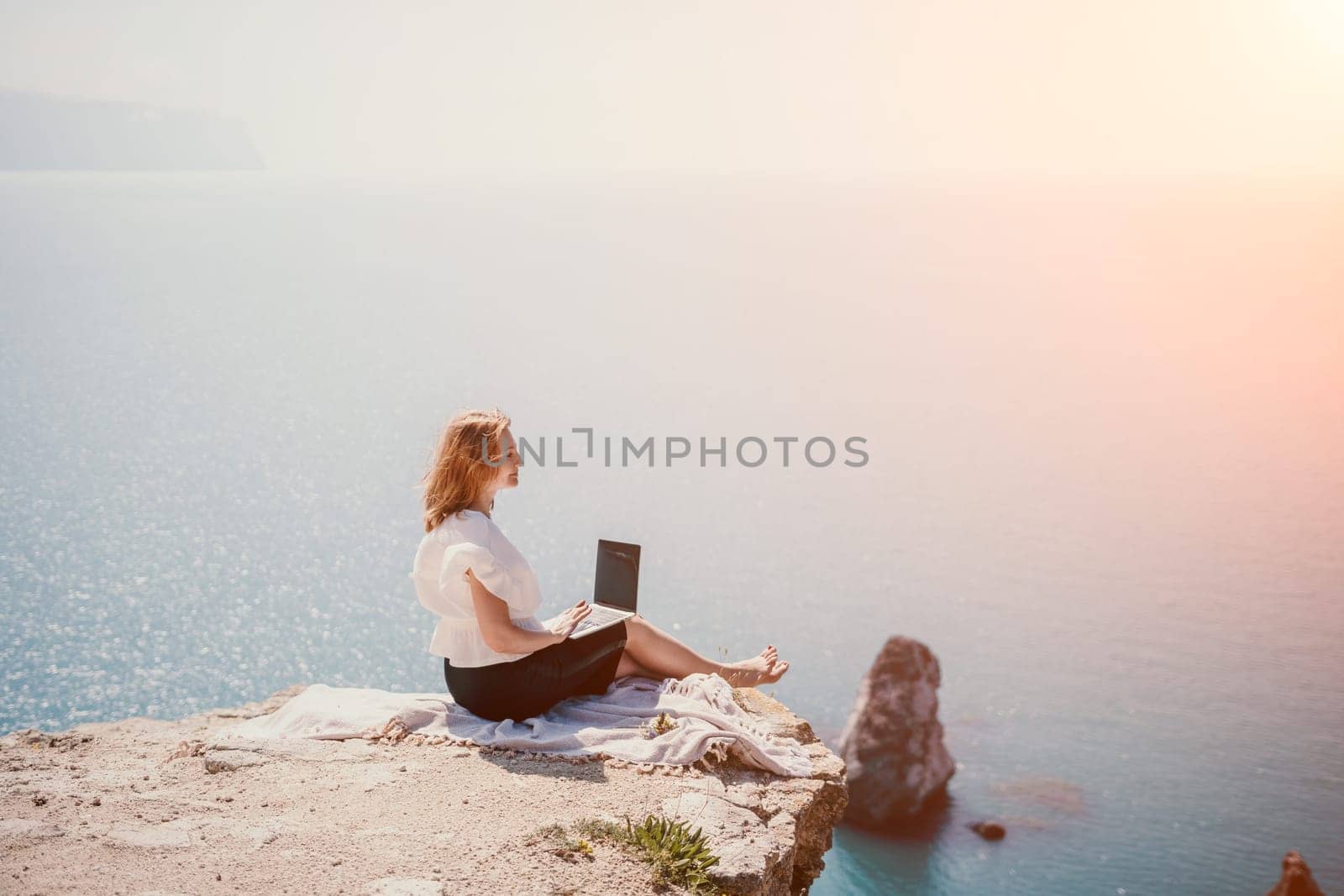 Successful business woman in yellow hat working on laptop by the sea. Pretty lady typing on computer at summer day outdoors. Freelance, travel and holidays concept.