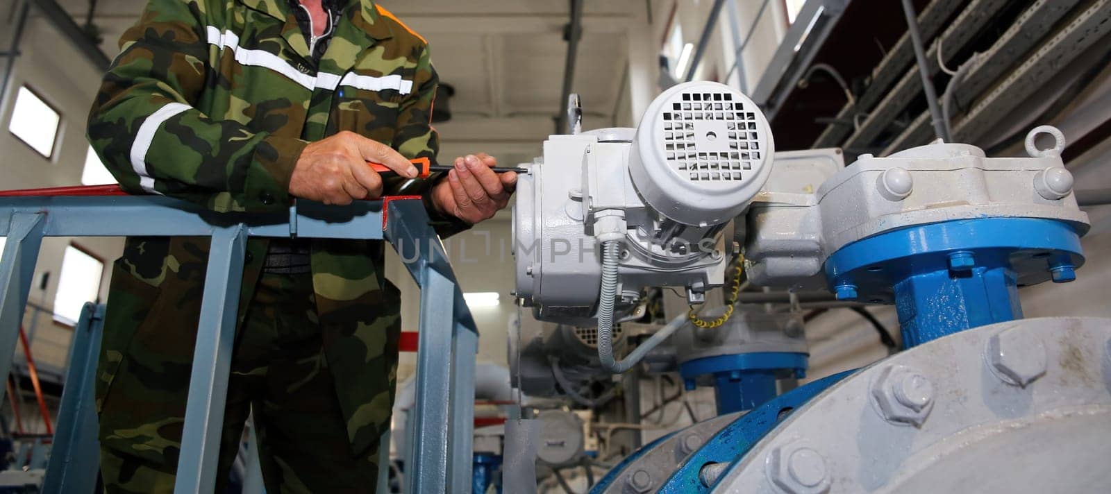 In a bustling factory setting, a skilled technician clad in a camouflage uniform diligently works on a large valve assembly. The atmosphere is filled with the hum of machinery and the scent of industrial lubricant.