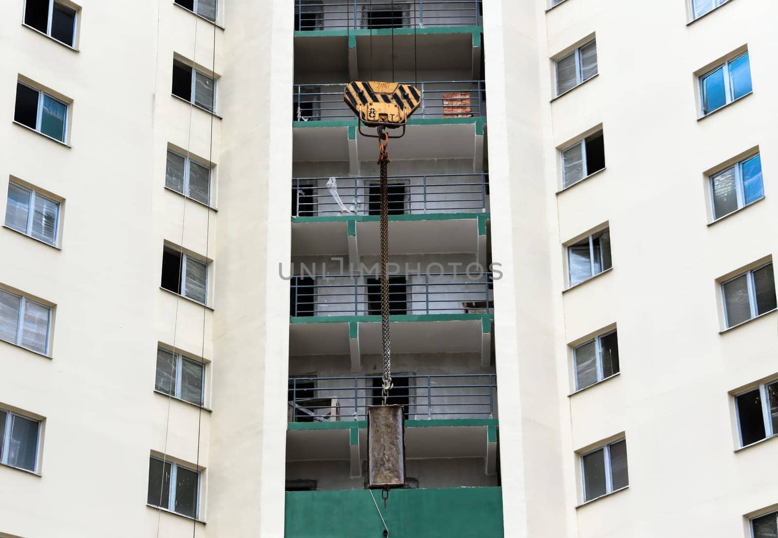 A towering crane descends from the sky, expertly maneuvering a heavy load toward the upper floors of a sleek apartment building. The structure features multiple balconies, with fresh paint adorning its walls. Workers below monitor the operation, ensuring safety and precision as the materials are lifted to the awaiting construction site.
