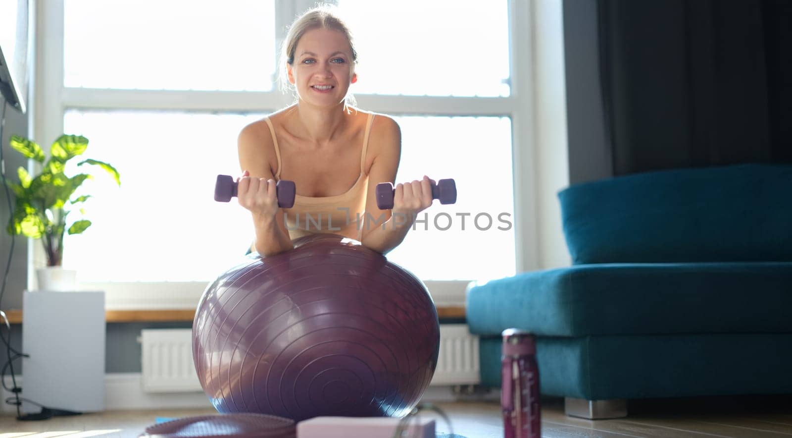 Woman doing exercises with dumbbells on a ball at home, sunny, close-up. Ball resistance training for women over 30