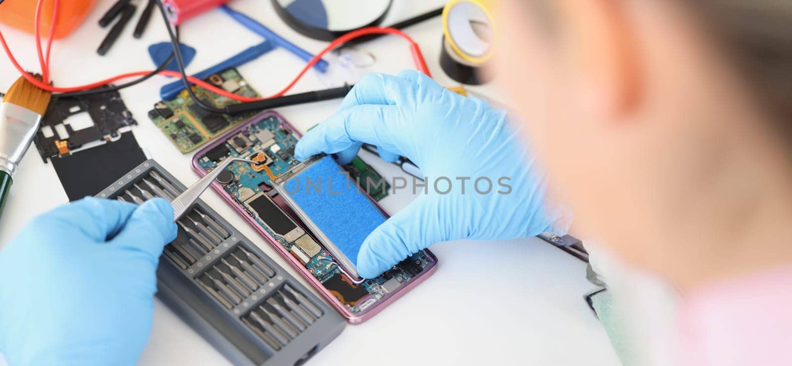Hands in blue gloves with tweezers are repairing the battery from a smartphone, close-up. Mobile phone repair service