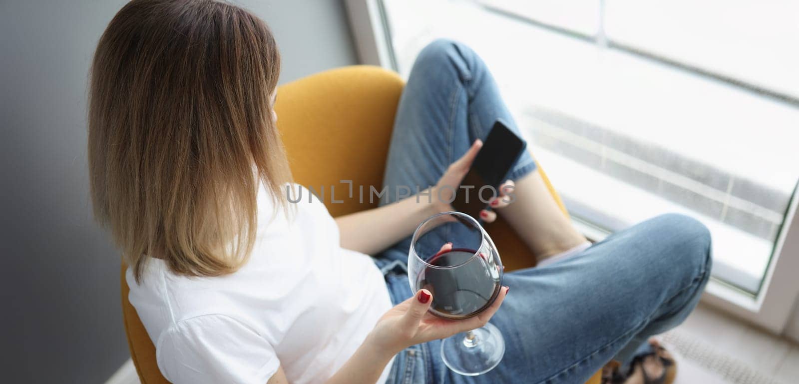 A woman at the window holds a glass of red wine and a smartphone, top view, close-up. Loneliness, stress relief, self-isolation