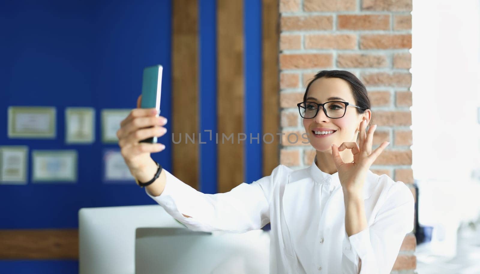 A woman in the office speaks via video calling on a smartphone, showing ok gesture close-up. Stylish businesswoman