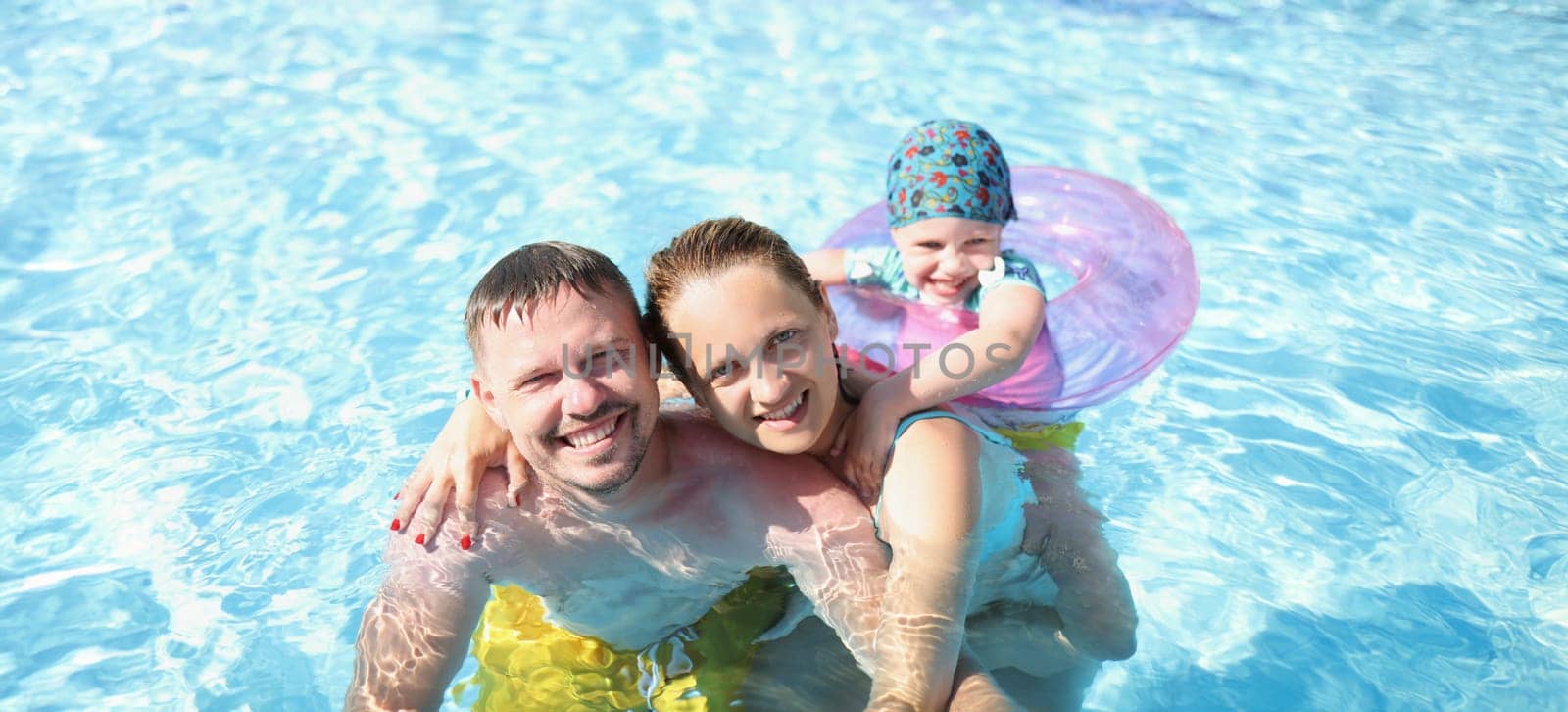 Husband with wife and child swim together in the pool, close-up. Happy family having fun in blue transparent water