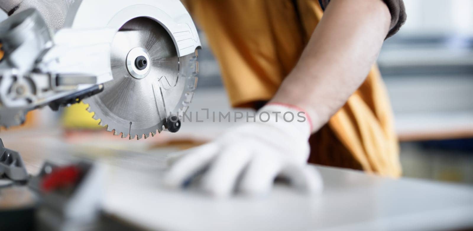 Male hands in gloves next to a circular saw, close-up, blurry. Woodworking workshop, sawing boards, carpenter equipment