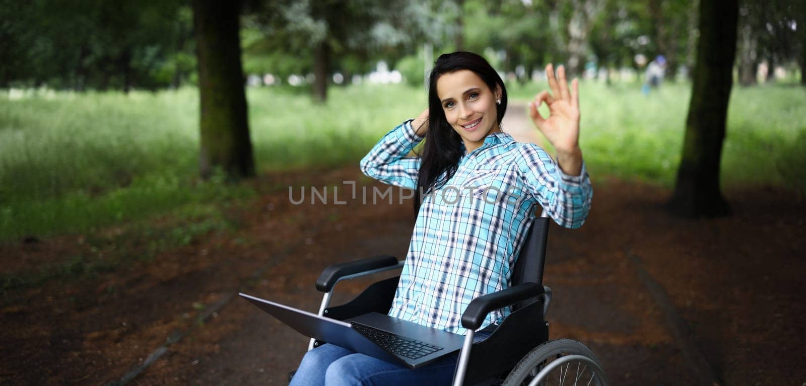 A young disabled woman sits in a wheelchair in the park with a laptop and shows the gesture ok. Psychological assistance after trauma