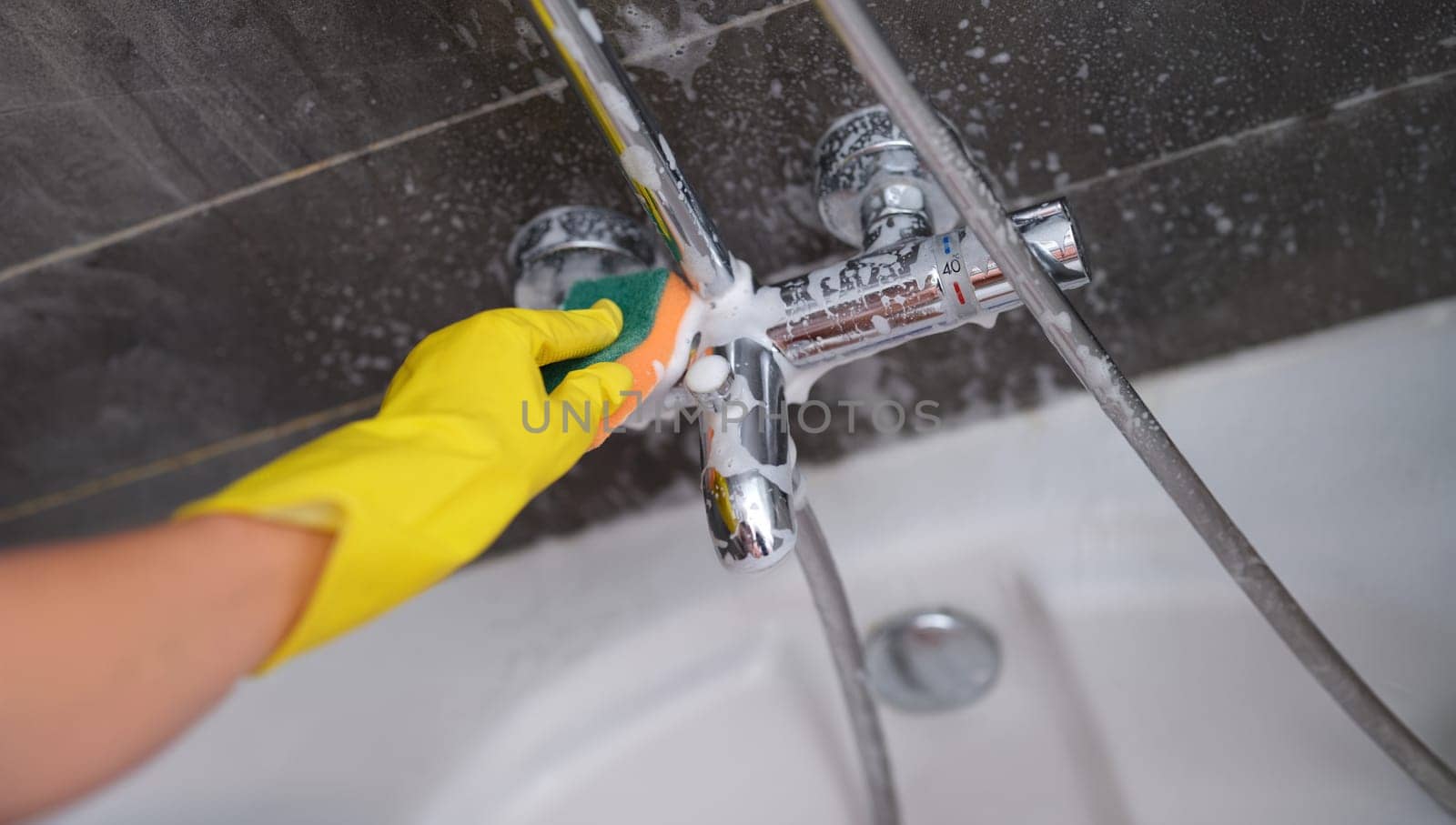 A hand in a yellow glove washes the mixer in the bathroom with a soapy sponge, close-up. Bathroom cleaning, disinfection