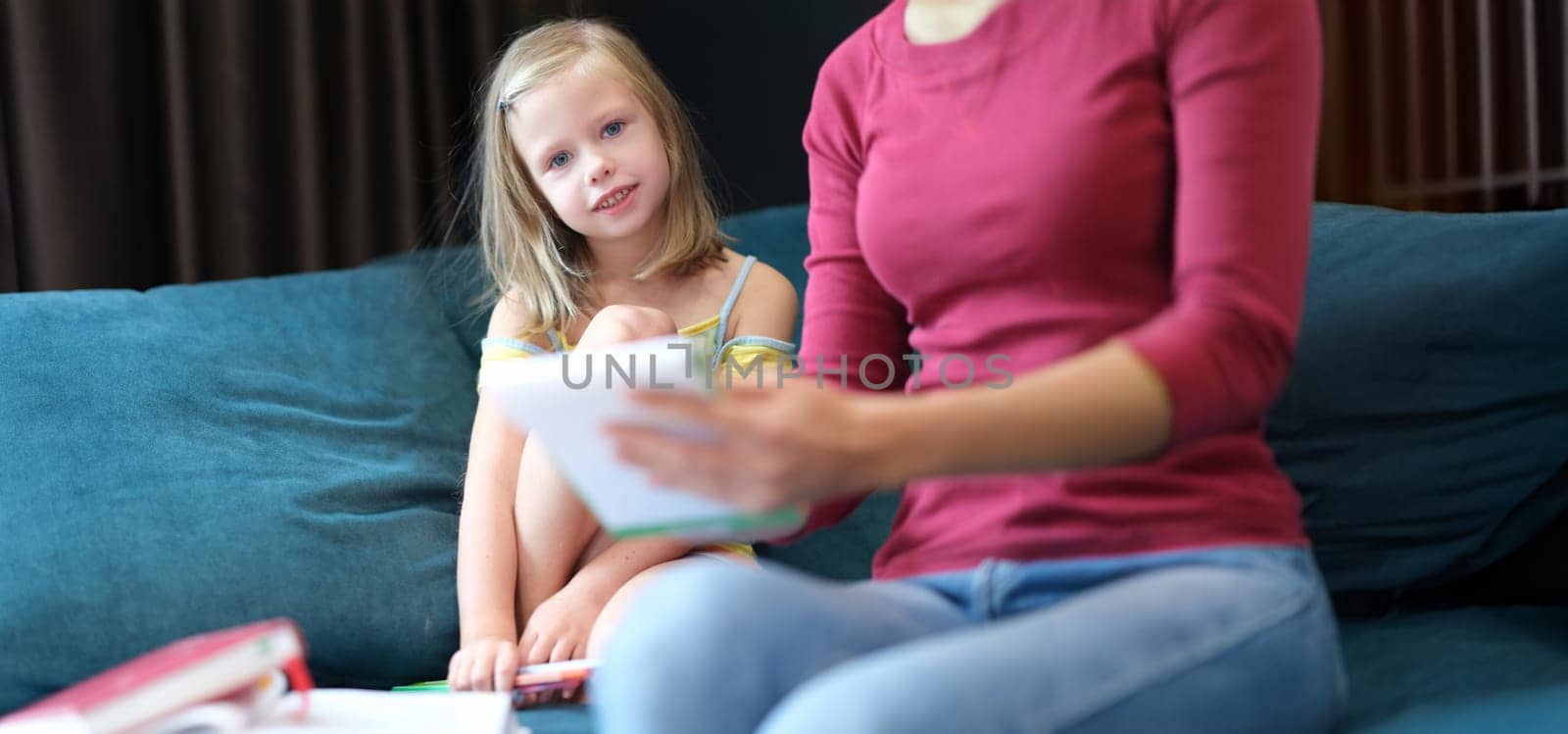 A little girl with her mother are sitting at home on the couch with books, close-up. Parent checks the child's school assignments
