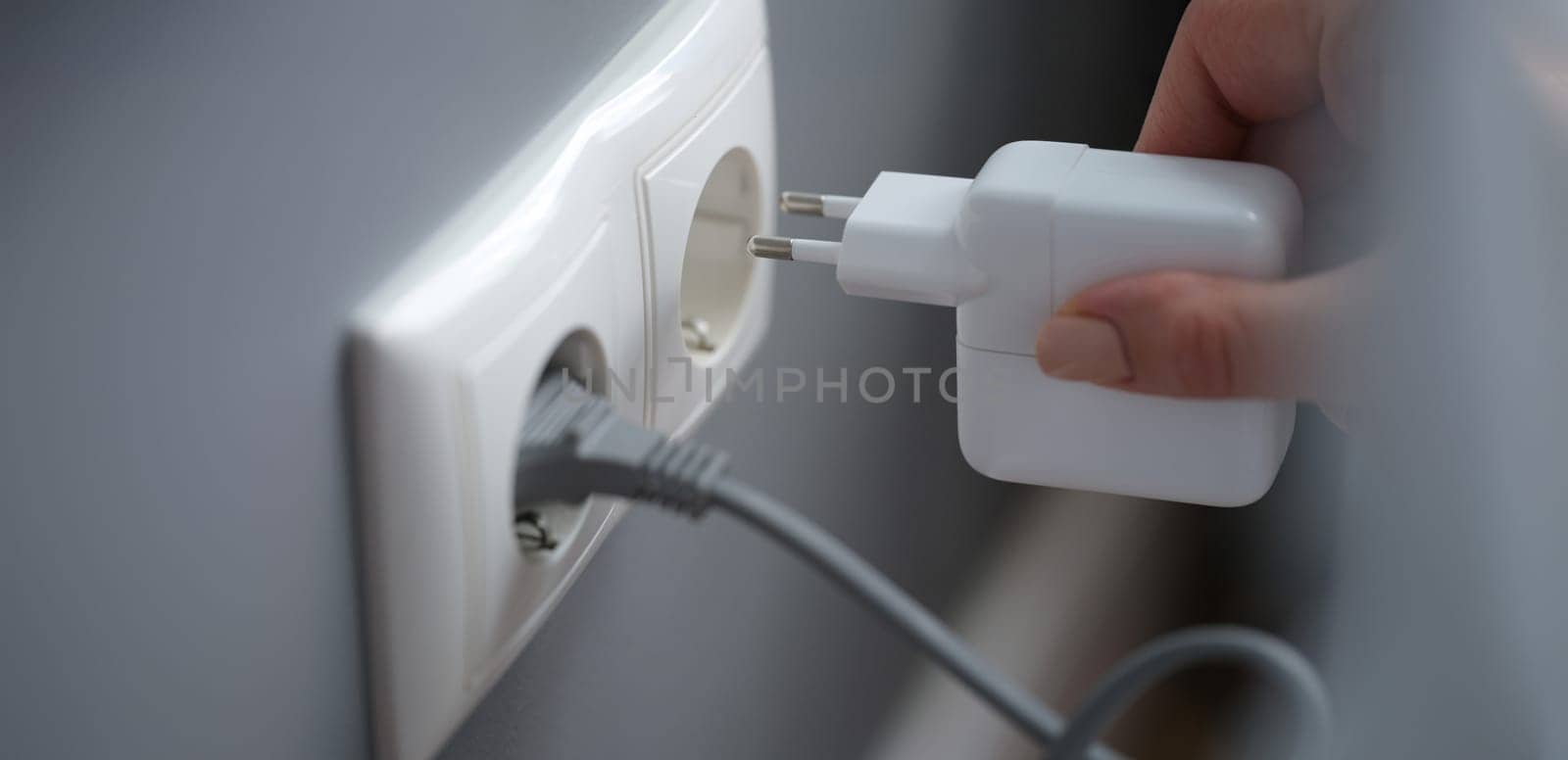 Female fingers plug the adapter into an outlet, close-up. White socket on gray wall background, electrician in apartment