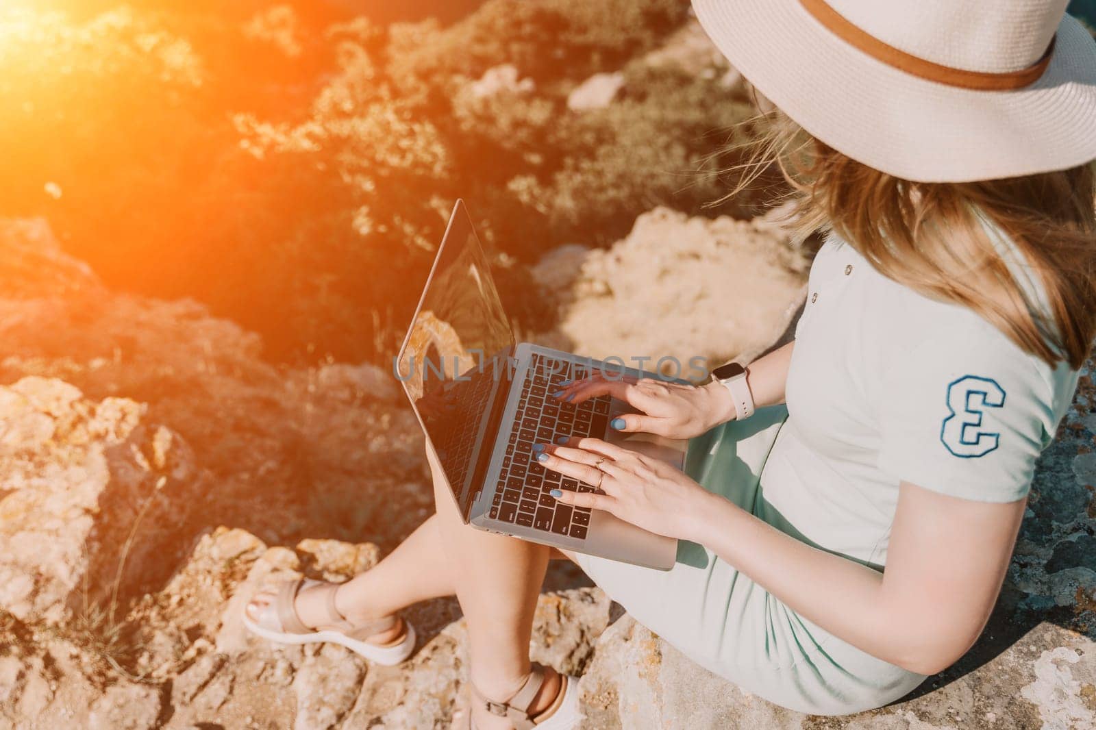 A woman in a white dress and hat sits on a rocky cliffside and uses a laptop, bathed in warm sunlight.