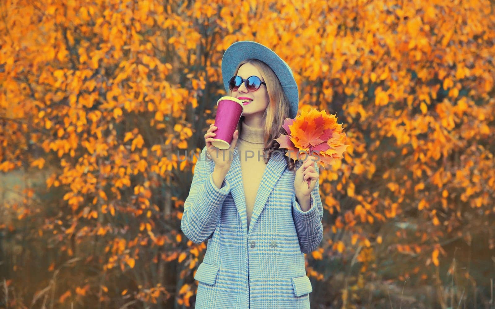 Autumn portrait of beautiful happy smiling woman with yellow maple leaves and cup of coffee wearing round hat and coat in the park