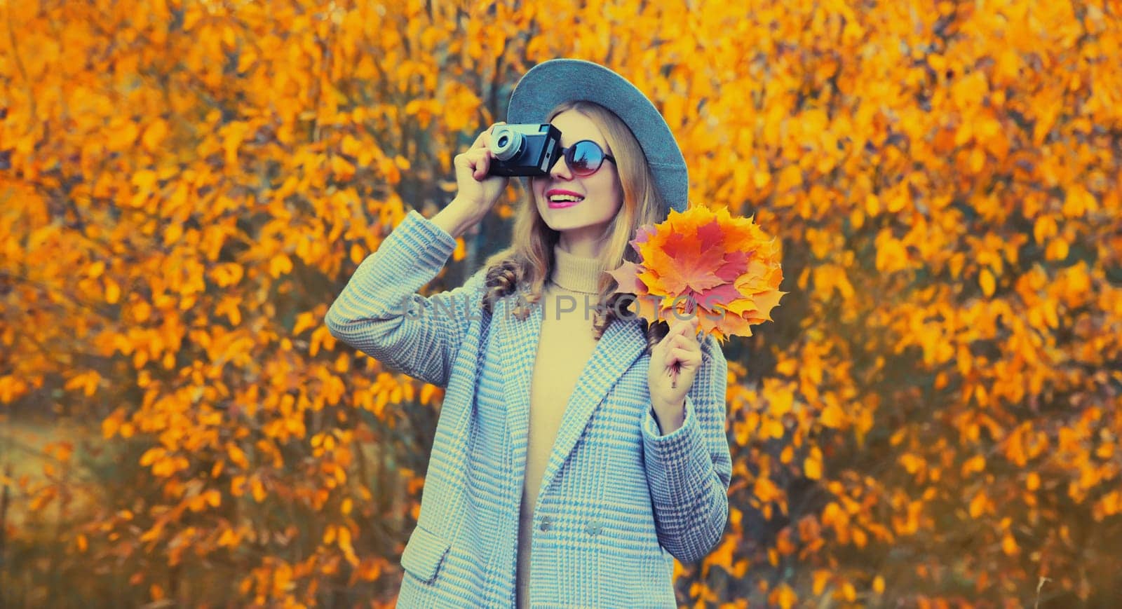 Autumn color style outfit, stylish young woman photographer with film camera and yellow maple leaves wearing round hat and coat in the park