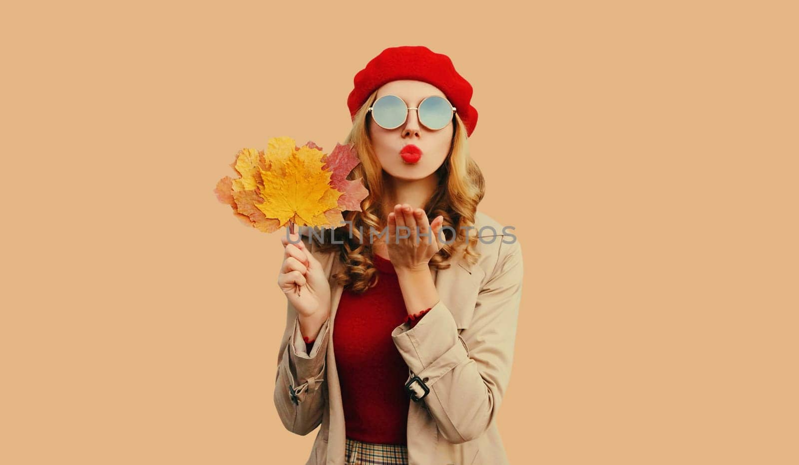 Autumn color style outfit, portrait of stylish beautiful woman model with yellow maple leaves blowing her lips sends sweet kiss wearing red french beret hat, round sunglasses on brown background