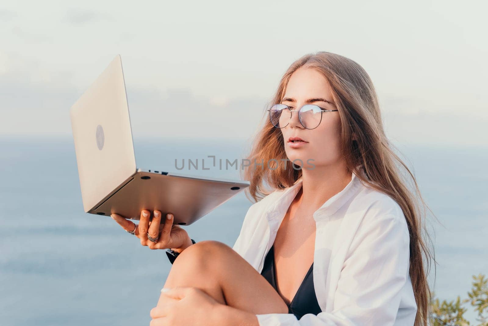 Digital nomad, woman in the hat, a business woman with a laptop sits on the rocks by the sea during sunset, makes a business transaction online from a distance. Freelance, remote work on vacation.
