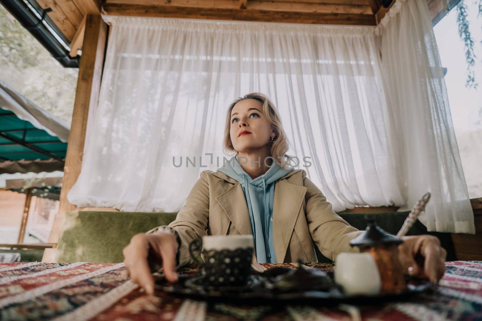 A woman sits at a table in a outdoor setting, enjoying a cup of Turkish coffee. The setting is cozy and inviting, with natural light filtering through the curtains.
