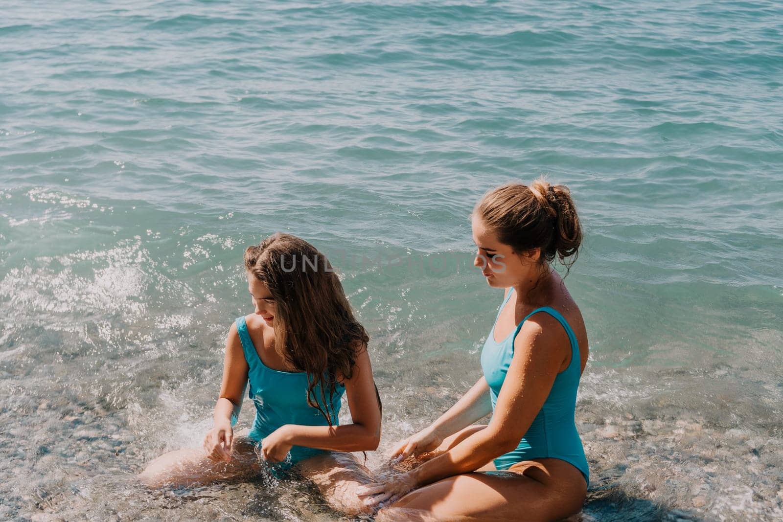 Two women in blue swimsuits are sitting in the shallow water on a pebble beach. The water is clear and blue.