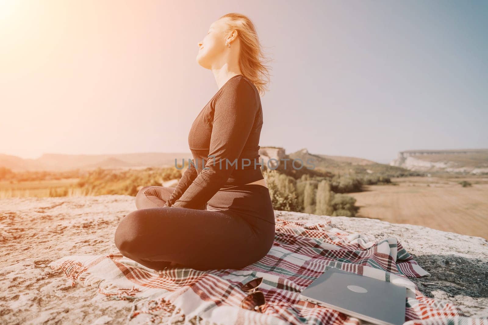 A woman in black clothing sits cross-legged on a mountaintop with a blanket and a laptop beside her, looking peaceful and serene.
