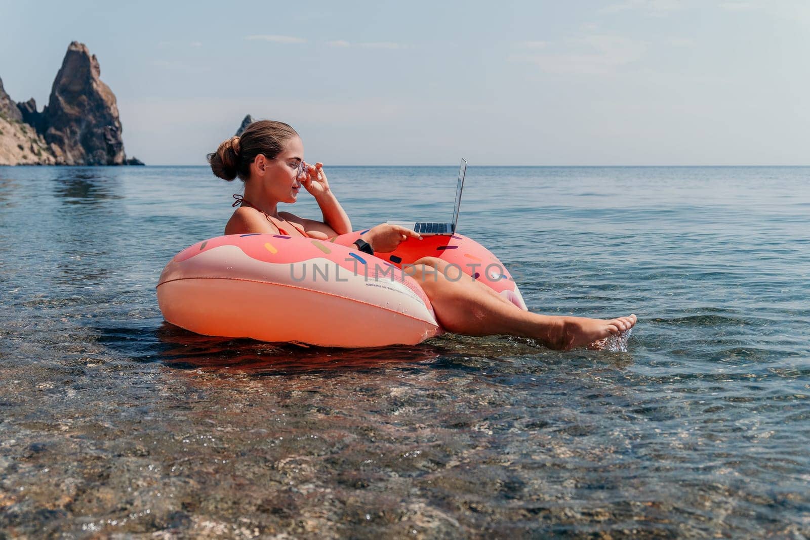 Woman freelancer works on laptop swimming in sea on pink inflatable ring. Pretty lady typing on computer while floating in the sea on inflatable donut at sunset. Freelance, remote work on vacation
