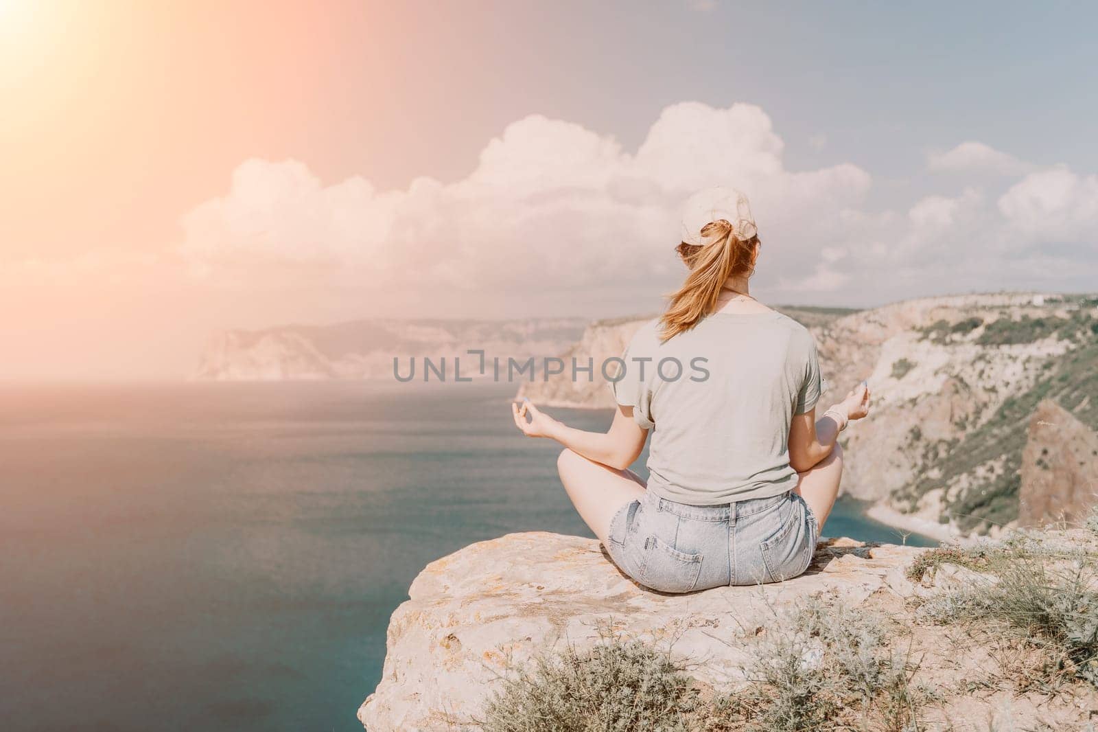 A woman sits in a yoga position on a rocky cliff overlooking a vast body of water, bathed in warm sunshine.