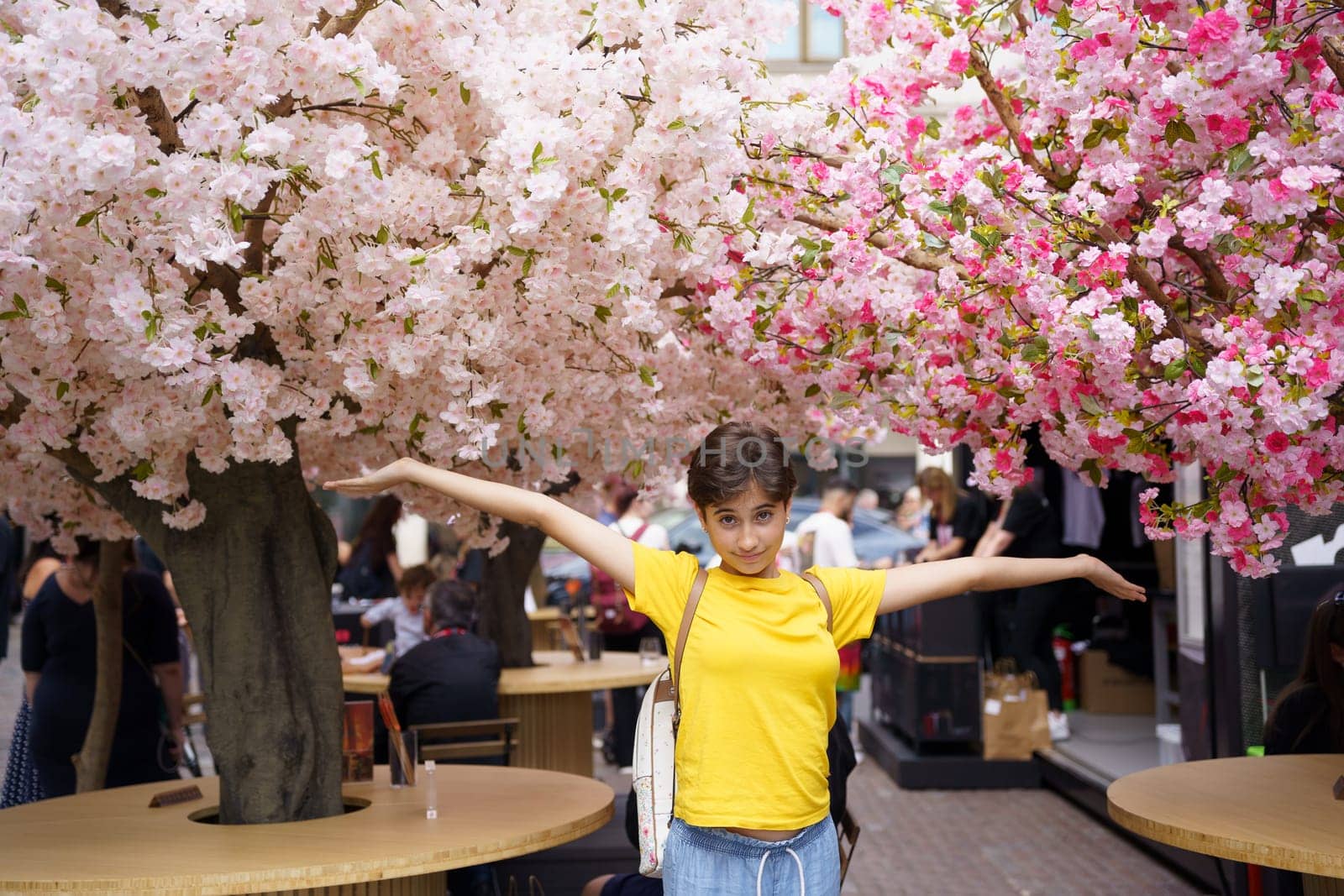 A cheerful young girl joyfully poses with her arms outstretched, amidst the stunningly beautiful cherry blossom trees that fill a lively and vibrant marketplace full of life and energy