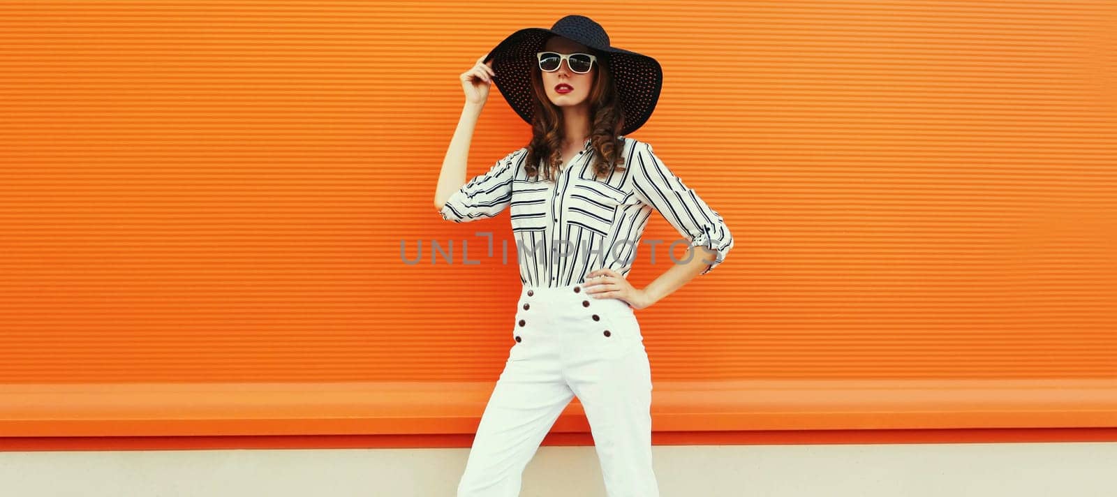 beautiful young woman posing wearing a white striped shirt, black round hat on orange background