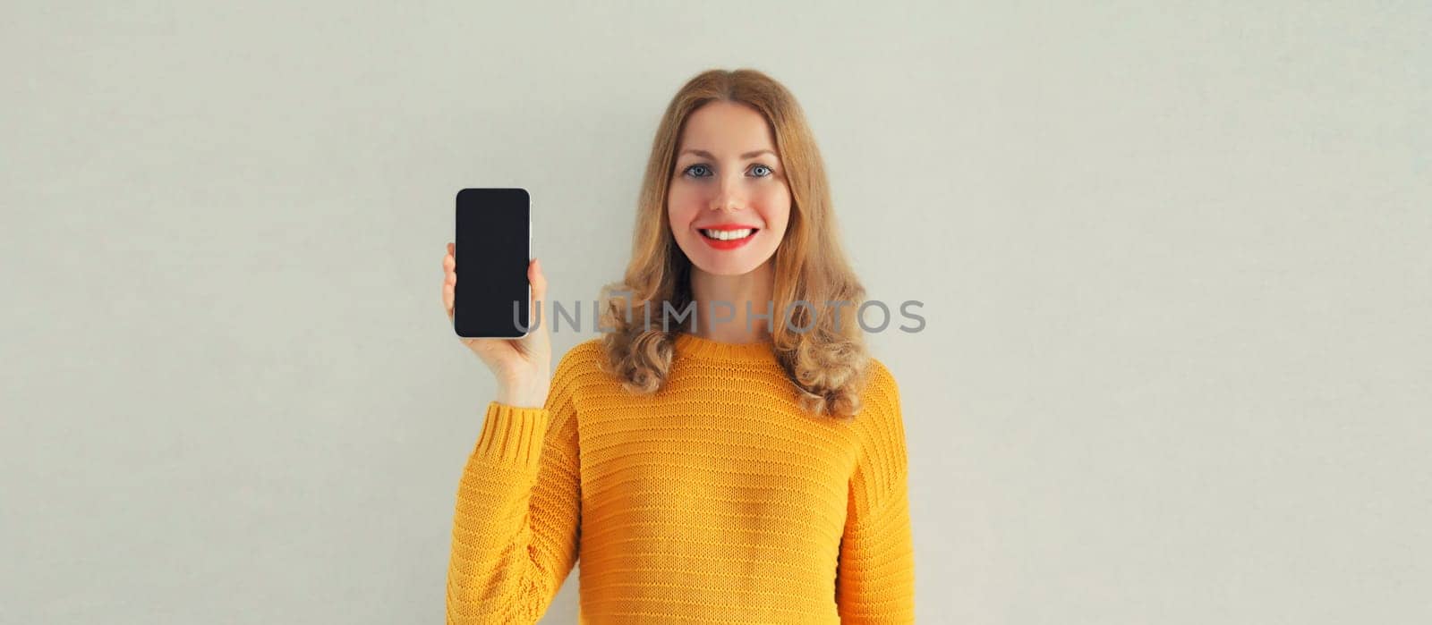 Portrait of happy smiling young woman showing black blank smartphone screen on gray background