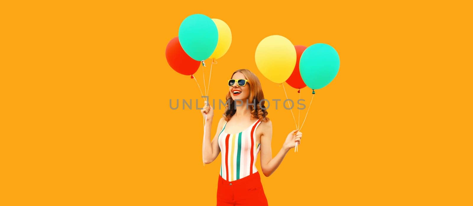 Summer portrait of happy smiling young woman with bunch of colorful red yellow balloons having fun wearing shorts, striped t-shirt, sunglasses on orange background