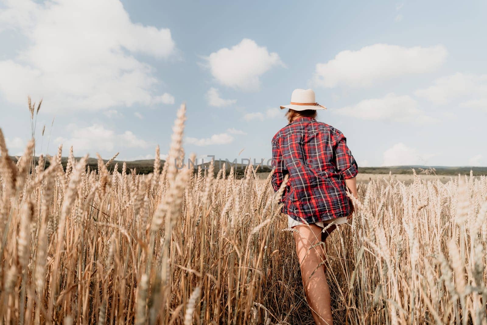 A woman in a plaid shirt and a hat walks through a field of wheat.