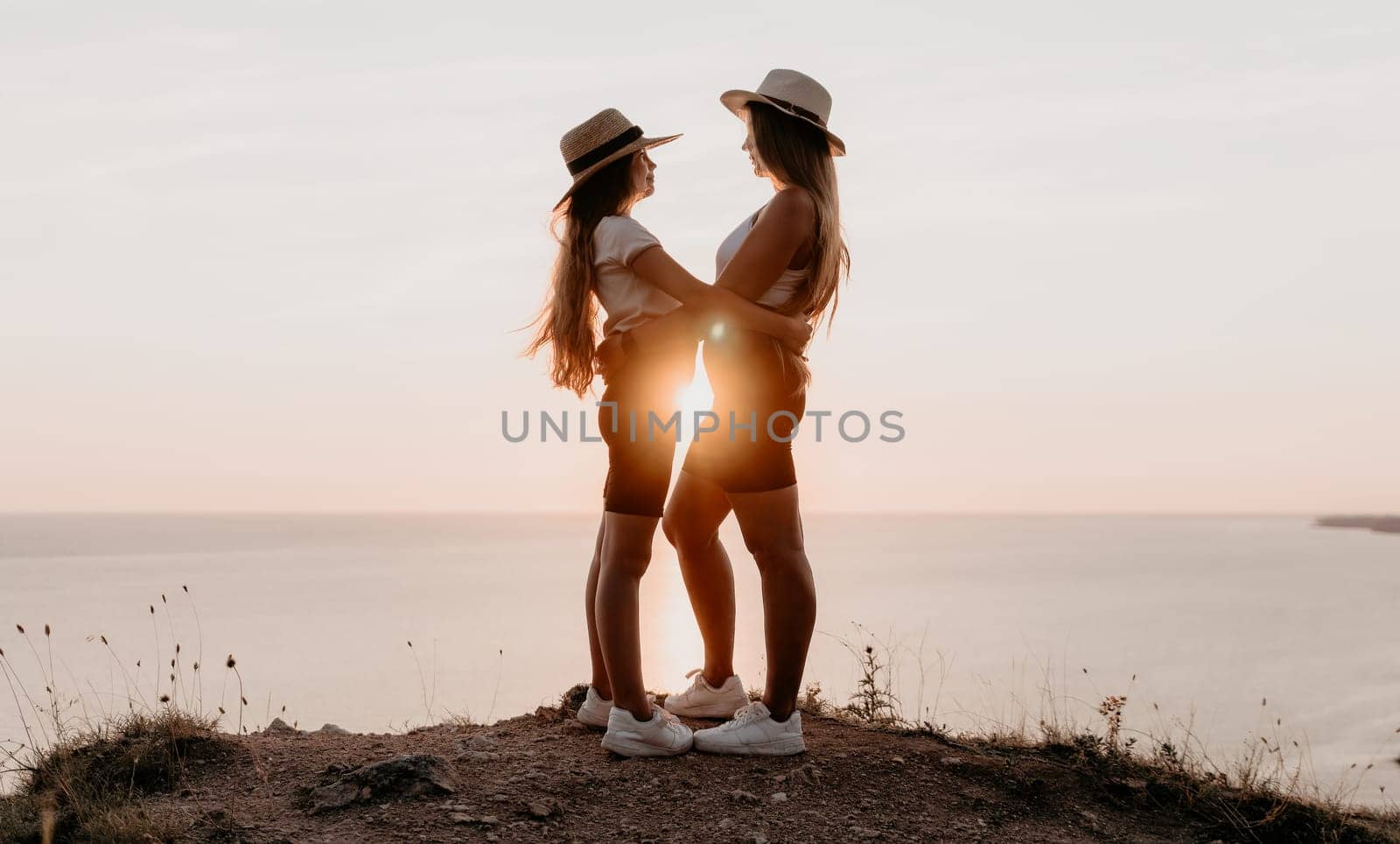 Two women in matching outfits and hats stand with arms around each other on a cliff overlooking the sea, bathed in the golden light of the setting sun.