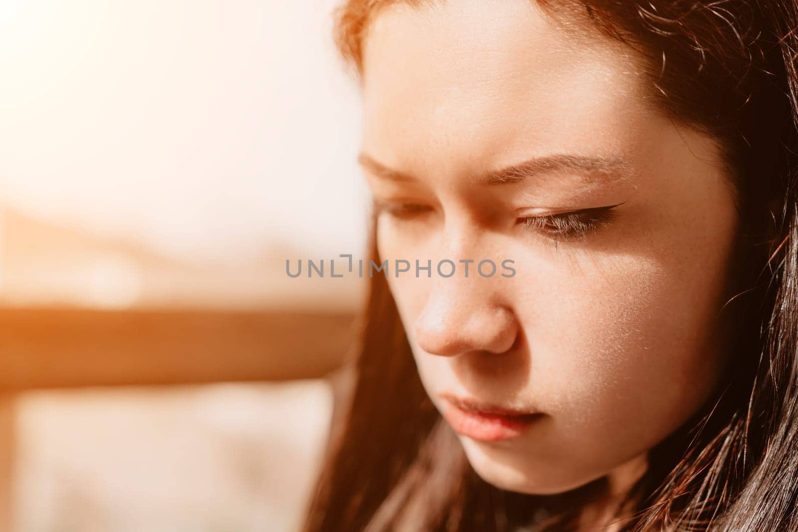 A close-up portrait of a young woman with long brown hair, gazing downward in a contemplative mood, bathed in warm sunlight.