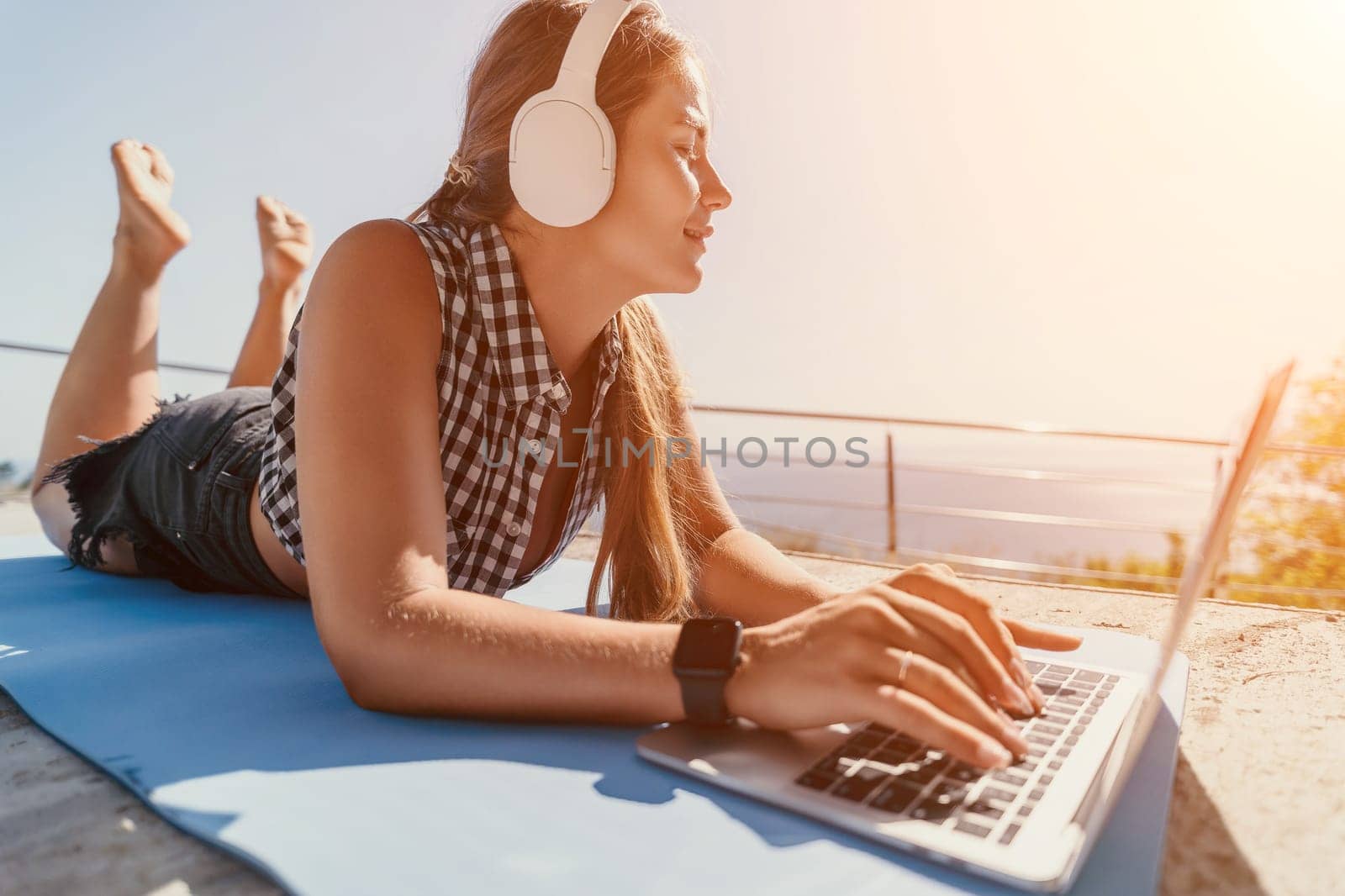 A young woman lies on a blue mat, enjoying the ocean view while using a laptop and listening to music with headphones.
