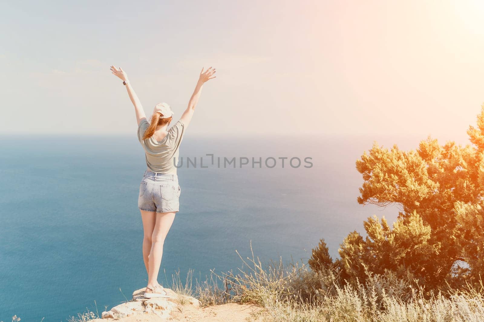 A woman with arms raised in triumph stands on a cliff overlooking a vast blue sea, with a sun-drenched sky above.